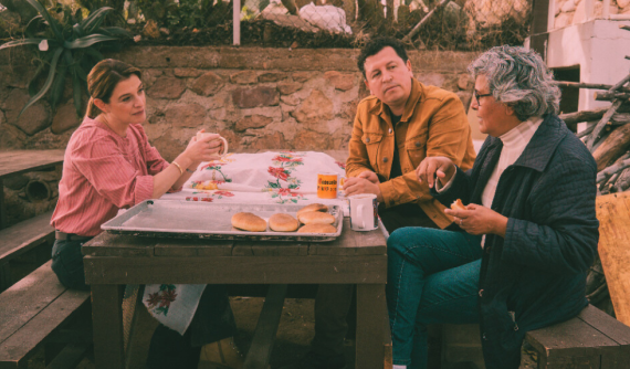 Image of activist and architect Ronald Rael wearing a brown jacket seated between Patti Jinich in a pink button down and blue jeans on the left and Angelica Macias in a black jacket with a white shirt and blue jeans on the right sharing fresh bread